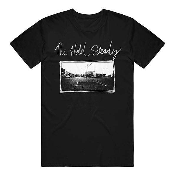 Black tee by The Hold Steady with the band’s name in white script above a black-and-white Minneapolis street scene, framed in a hand-drawn style—a tribute to their iconic Separation Sunday era.